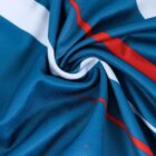 A close-up of a twisted fabric with blue, white, and red colors creating a spiral pattern. The fabric has a smooth texture with small red dots scattered across the blue areas.