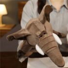 A person holding a plush elephant hand puppet. The brown elephant has large ears, a trunk, tusks, and detailed stitching. A lamp and a blurred background with a piece of furniture are visible.