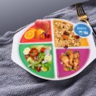 A divided plate features an apple and citrus fruits, granola with nuts and berries, a salad with greens and eggs, and fried chicken. A label shows a dairy icon. The plate is on a textured gray surface with a fork nearby.