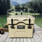 A beige cooler with a black handle sits on a wooden deck, featuring the brand name Cumbria. In the background, there is a blurred view of a lake and forested mountains. Nearby, there are glasses, fishing gear, and a fishing rod.
