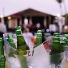 A clear ice bucket filled with green glass beer bottles, some of which have condensation, sits on a table. Blurred in the background is a stage with people and lights, suggesting an outdoor event or concert.