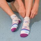 A person sitting on a beige stool wears socks featuring a pattern of loops in purple and blue. The socks have purple toe caps and heels, with a blue and purple stripe and the word akaden near the toes, on a blue speckled floor.