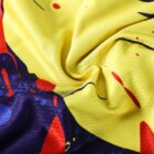 Close-up of a vibrant fabric with a swirling pattern. The design features splashes and strokes of red, blue, and dark colors on a bright yellow background, creating an abstract, dynamic effect.