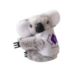 Stuffed koala toy with fluffy gray fur and a black nose, wearing a white bib with a purple logo and the word BRAND on it. The koala is holding a small black plastic fork.