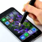 A hand using a stylus on a smartphone screen, displaying a vivid purple flower wallpaper. Several app icons are visible, including Mail, Calendar, Photos, and Music, arranged along the bottom of the screen.