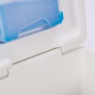An open white cooler with a blue ice pack visible inside the lid. The ice pack has some embossed symbols on it. The cooler is positioned on a light surface.