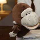 A person is using a soft, brown and gray monkey hand puppet with a smiling expression. The background features a blurred lamp and indoor setting.