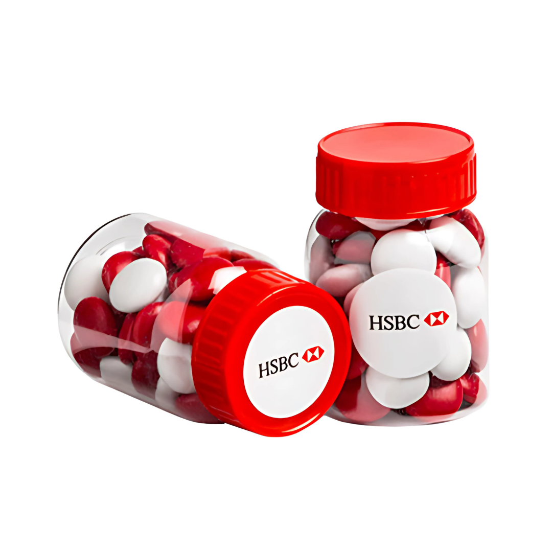 Two clear screw-top jars labeled HSBC, each filled with red and white capsule-shaped chocolate candies, feature red lids. One jar stands upright while the other lies on its side, each weighing 50g and intended for babies.