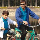 A man and a boy are casually dressed and riding bicycles near a pond. The man wears sunglasses, a blue jacket, and a teal shirt, while the boy has a white jacket and blue shirt. Palm trees are visible in the background, with BOCINI CASUAL text on the image.