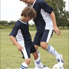 A man and a boy in matching navy and white soccer uniforms playfully interact on a grassy field with a soccer ball. Both are smiling, and the scene conveys a sense of joy and sportsmanship. Trees are visible in the background.