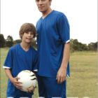 A man and a boy, both wearing blue soccer jerseys, stand on a grassy field. The boy holds a soccer ball. The image has the word SPORTS at the bottom and BOCINI at the top left corner.
