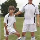 A man and a boy are standing on a grassy field wearing matching white sports jerseys and shorts with black and blue accents. The boy is resting his foot on a soccer ball. BOCINI and SPORTS are written on the image. They are smiling.