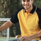 A person wearing a yellow and black polo shirt sits on a wooden bench outdoors, smiling. The word CASUAL is written at the bottom, and the logo BOCINI appears at the top left. A blurred background suggests a setting near a tennis court.