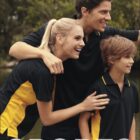 Three people outdoors, wearing matching black and yellow sports polo shirts, smile and look into the distance. The background is a natural setting with trees. The word SPORTS is prominently displayed at the bottom.