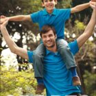 A man wearing a blue polo shirt carries a smiling boy on his shoulders, both wearing blue jeans. They are outdoors with trees in the background. The word SPORTS is at the bottom, and BOCINI is at the top left corner.