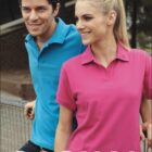 Man in a blue polo shirt and woman in a pink polo shirt smiling and leaning against a railing outdoors. The word CASUAL is displayed at the bottom. Trees and a brick building are in the background.