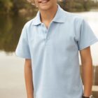 A smiling child wearing a light blue polo shirt stands outdoors near a lake. Trees and water are visible in the background.
