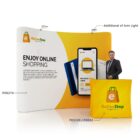 Exhibition booth display with the logo Online Shop, featuring a slogan, a mobile shopping app image, and a price tag. A man in formal attire stands next to the booth. A branded table cover is in front, and theres an additional light on top.