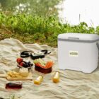 A picnic setup on a striped blanket near a river. Items include a white cooler, vegetables, apples, grapes, chips, dips, condiments, a camera, and a small jar of honey, all surrounded by lush greenery.