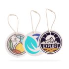 Three round air fresheners with strings. The first has colorful abstract shapes, the second features a turquoise leaf design, and the third shows mountains with the word EXPLORE below.