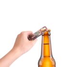 A hand uses a bottle opener to remove the cap from a brown beer bottle. The background is plain white.