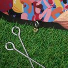 Close-up of two metal pegs on green artificial grass, next to a colorful illustration of people playing sports, with visible leaves in the top right corner. A black edge with a loop and golden clasp is at the top.
