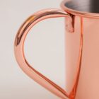 Close-up of the handle of a copper mug against a light beige background. The handle is smooth and shiny, reflecting light. The mug interior appears to be made of stainless steel.
