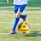 A person wearing a white shirt, blue shorts, and blue socks is playing on a grassy field, kicking a colorful yellow soccer ball with printed graphics and Telesale written on it.