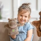 A young girl with a tiara smiles while playing with a hippo hand puppet. She is sitting on a sofa, and the setting appears to be indoors with a blurred background.