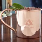Copper mug filled with ice and garnished with a lime wedge, placed on a wooden table. The mug features a logo with a geometric star design and the words BRAND NAME.