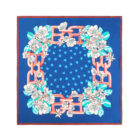A blue square scarf featuring a floral pattern with white flowers and green leaves. Red chain-like designs frame the flowers, and small blue floral motifs are scattered in the center.