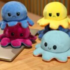 Four plush octopus toys are placed on a wooden surface and a laptop. Each has a different side showing: two have sad expressions in blue and pink, while the others display happy faces in yellow and blue.