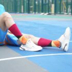 Person sitting on a colorful basketball court wearing white sneakers, bright red and orange argyle socks, and blue and orange athletic shorts. Their legs are extended forward, showing off the bold sock pattern.