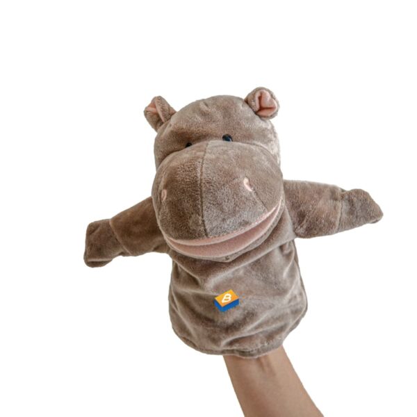 A hand holding a plush hippo hand puppet. The puppet is brown with pink details on its ears and nostrils. It has a big smile and blue eyes. Theres a small blue and orange label on its body. The background is plain white.