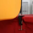 Close-up of a clamp securing an orange object to a red surface. The clamp is fastened tightly with a visible screw mechanism. The background is blurred, containing vertical lines that may be part of a structure or railing.