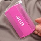 A person holds a small pink double-sided comb, packaged in clear plastic. The comb features two different sets of fine teeth and has text printed on it. The background is a textured gray fabric.