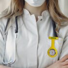 A healthcare professional with long hair, wearing a white coat and a medical mask, stands with arms crossed. A stethoscope hangs around their neck, and a yellow watch labeled CAIXA is attached to their chest pocket.