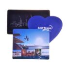 Three colorful mousepads: one heart-shaped, blue with SoftTrans text; another rectangular with a city skyline; and the last showing a vibrant seaside scene and small photos at the bottom.