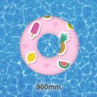 A pink inflatable swim ring with colorful fruit and ice cream designs, including a pineapple, lemon, popsicle, and watermelon, floats on clear blue pool water. 900mm is written below the ring.