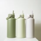 Three reusable water bottles in varying shades of green and off-white are lined up against a neutral background. Each bottle has a handle and a spout lid.