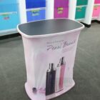A portable display stand with a black top and curved sides wrapped in a pink fabric featuring images of two perfume bottles and the text Black & Pink series Pearl Brand. Colorful product shelves are visible in the background.