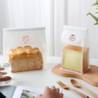 Two types of bread are in transparent, resealable bags on a table: a round bun loaf marked Gelato and a square loaf labeled 100%. A person leafs through a magazine, with a glass of water and plant visible.