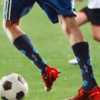 Close-up of a soccer player in blue shorts and branded navy socks dribbling a black and white soccer ball on a grassy field. The players red cleats are visible, with another player in the background in white shorts and red socks.