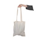 A persons hand holds a beige fabric tote bag by the straps. The person is wearing a black and white striped sleeve. The background is plain white.