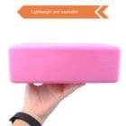 A hand holding a pink yoga block against a white background. The block is labeled as lightweight and washable in an orange graphic above. The person wears a black wristband.