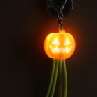 A glowing orange jack-o-lantern keychain with a smiling face and jagged teeth is attached to a clip. Long green strings dangle below, set against a dark background.
