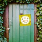 A green wooden door surrounded by green ivy. A sign with a yellow background hangs on the door, featuring illustrations of flowers and the words Spring Party. A bolt and padlock are installed on the doorframe.