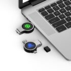 A laptop on a table with two USB security keys nearby. One key features a glowing green emblem, while the other displays a blue emblem with a triangular design. A USB connector cap is also visible beside them.