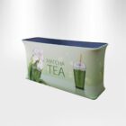A rectangular table with a printed fabric cover features images of matcha tea. The design includes a splash of green tea and a cup of iced matcha with a transparent lid. Text reads MATCHA TEA. The table surface is dark.