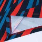 Close-up of a fabric corner with a bold geometric pattern in red, blue, and black. The fabric is folded to show stitching and the lighter underside. The design features elongated, angular shapes creating a dynamic, zigzag effect.